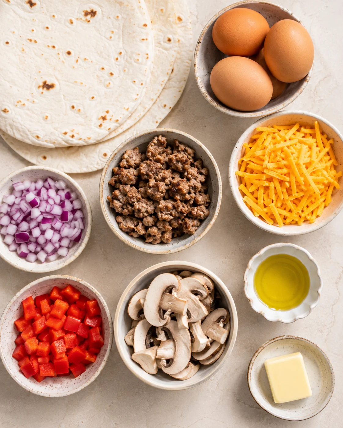 Top-down arrangement of tortillas, eggs, cooked sausage, shredded cheese, mushrooms, red pepper, onion, oil and butter
