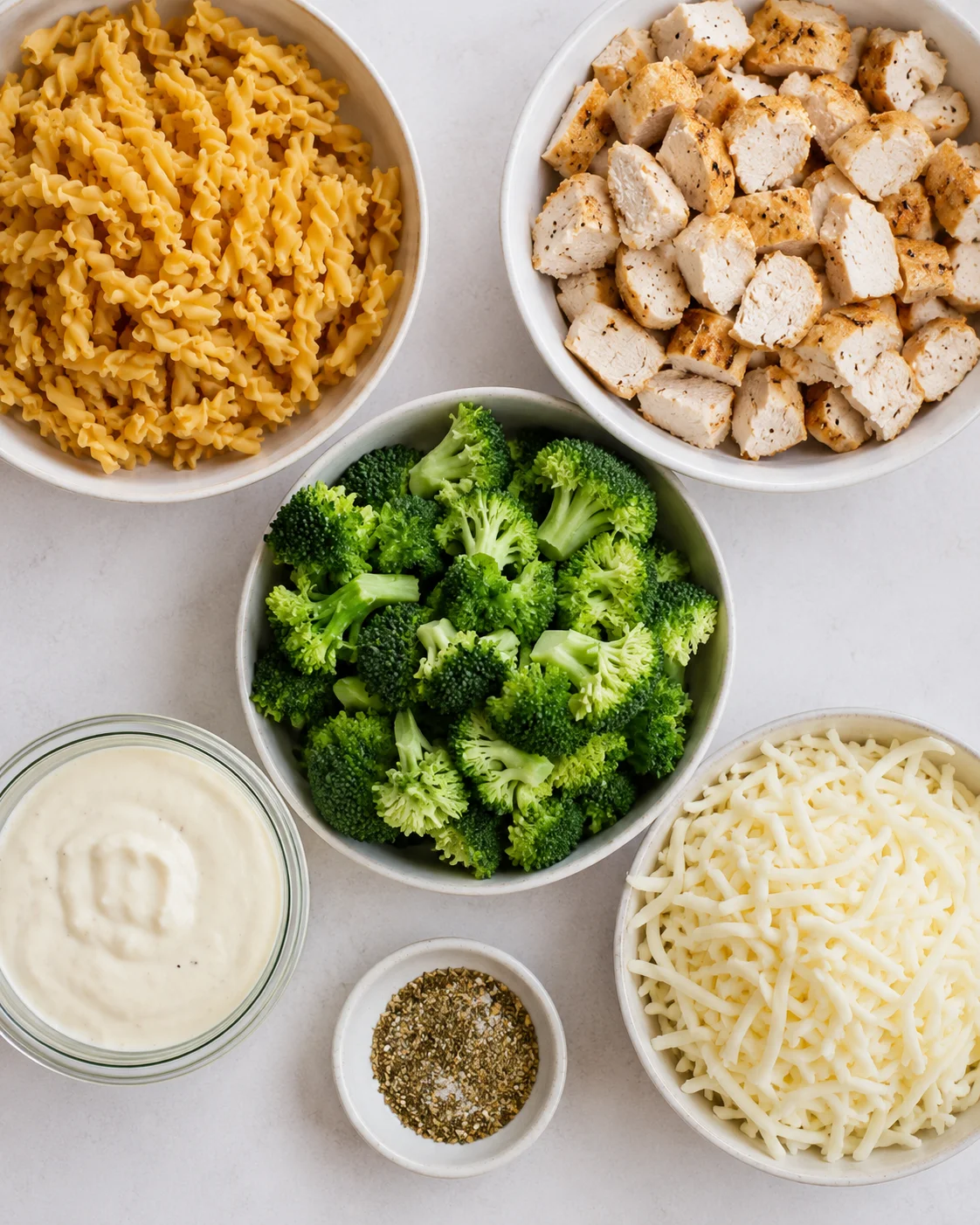 Top-down view of bowls with uncooked chickpea pasta, cooked diced chicken, broccoli, alfredo sauce, shredded mozzarella and seasonings