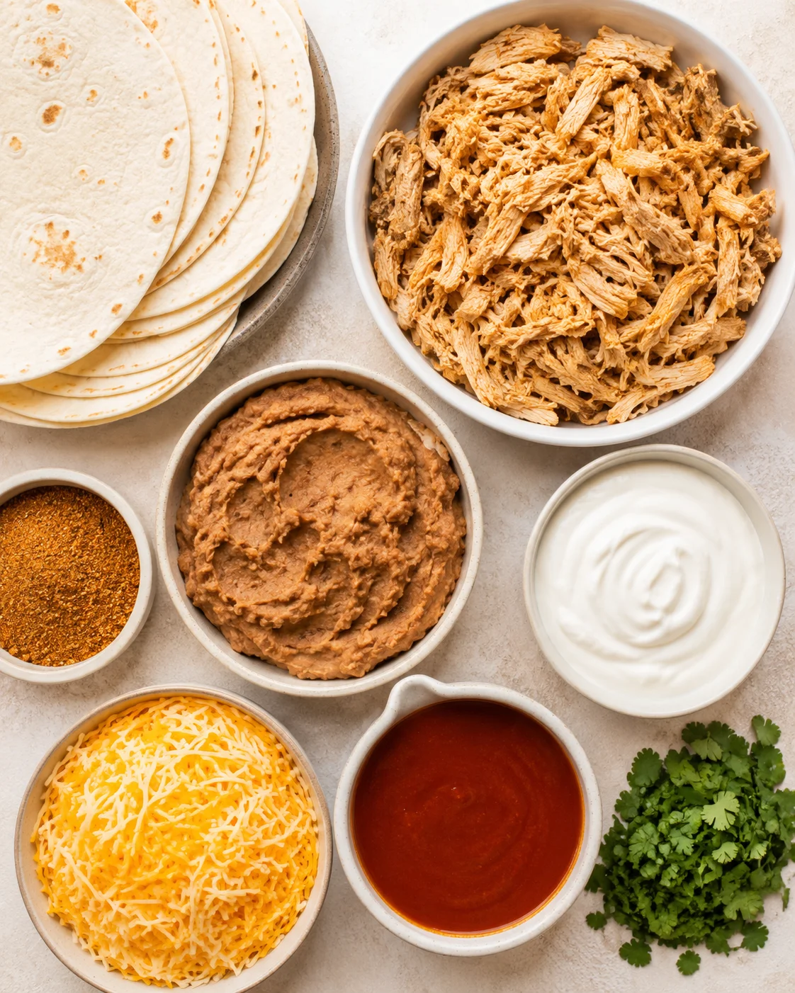 Top-down view of ingredients: tortillas, shredded chicken, refried beans, Greek yogurt, cheese, enchilada sauce, taco seasoning and cilantro