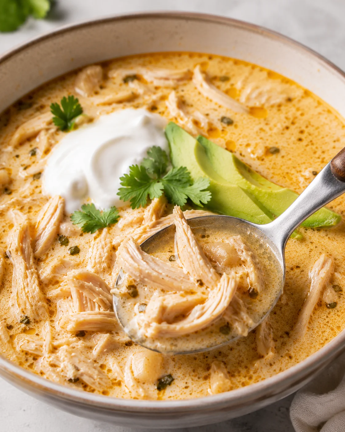 Keto white chicken chili in a bowl with shredded chicken, avocado slices, sour cream, cilantro and a spoon lifting a bite