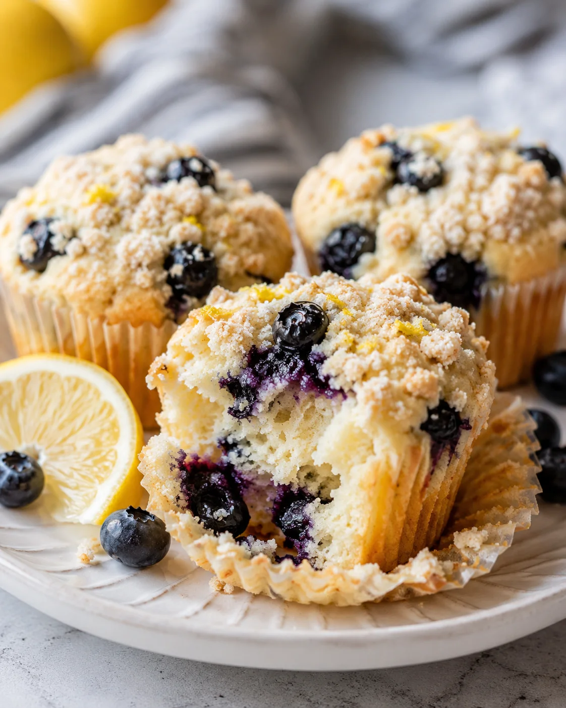 Plate with a partially eaten lemon blueberry muffin showing the interior crumb, with a lemon slice and extra blueberries beside it