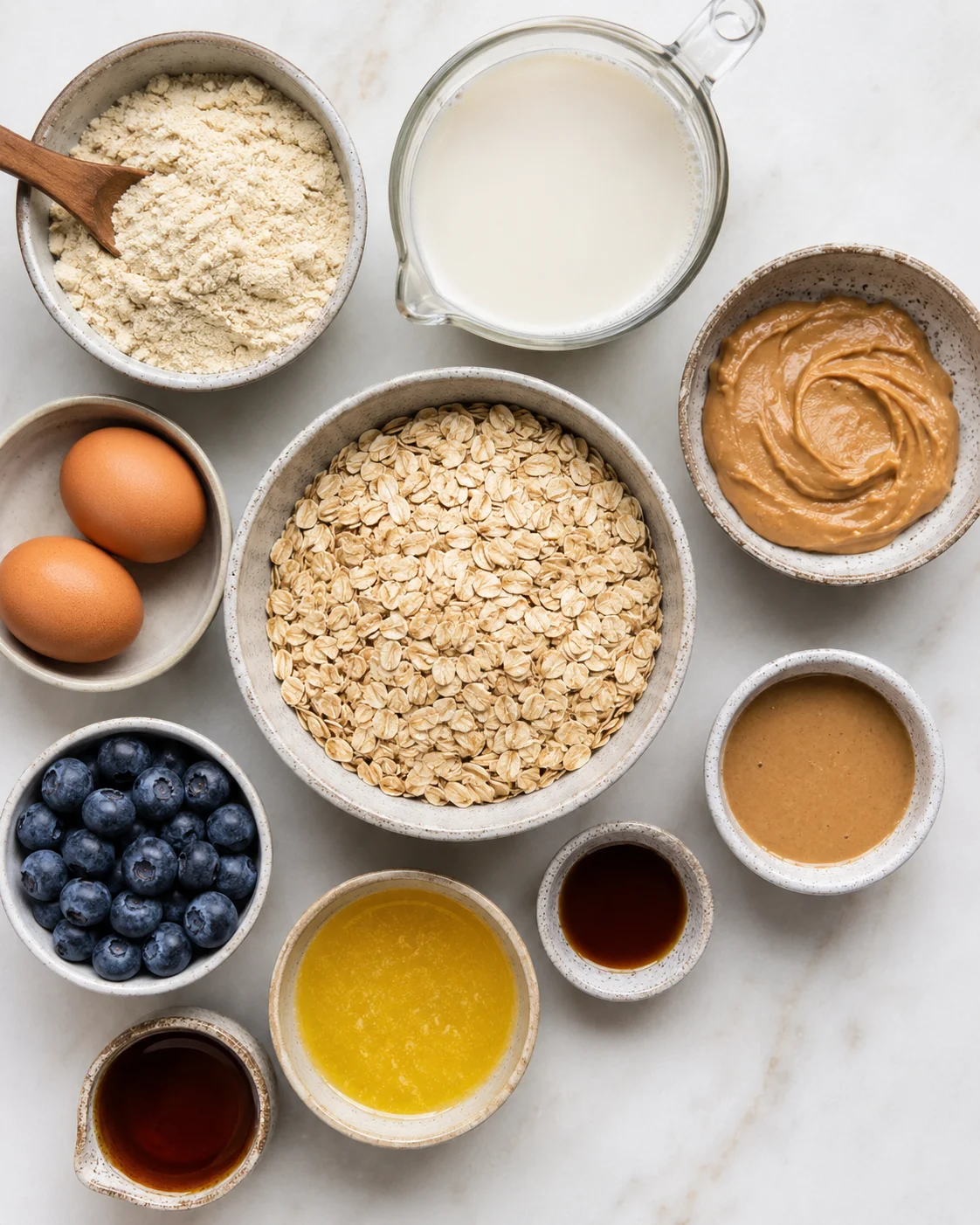 overhead view of a central bowl of oats surrounded by flour, milk, eggs, blueberries, and peanut butter.