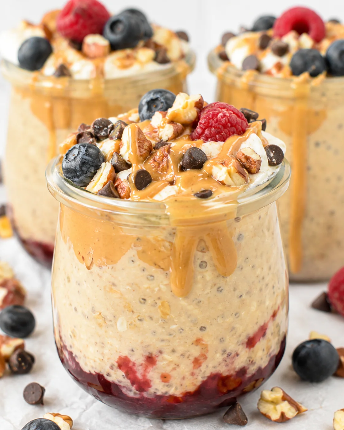 glass jar of overnight oats with berry layers, peanut butter drizzle, blueberries, raspberries, and chopped nuts.