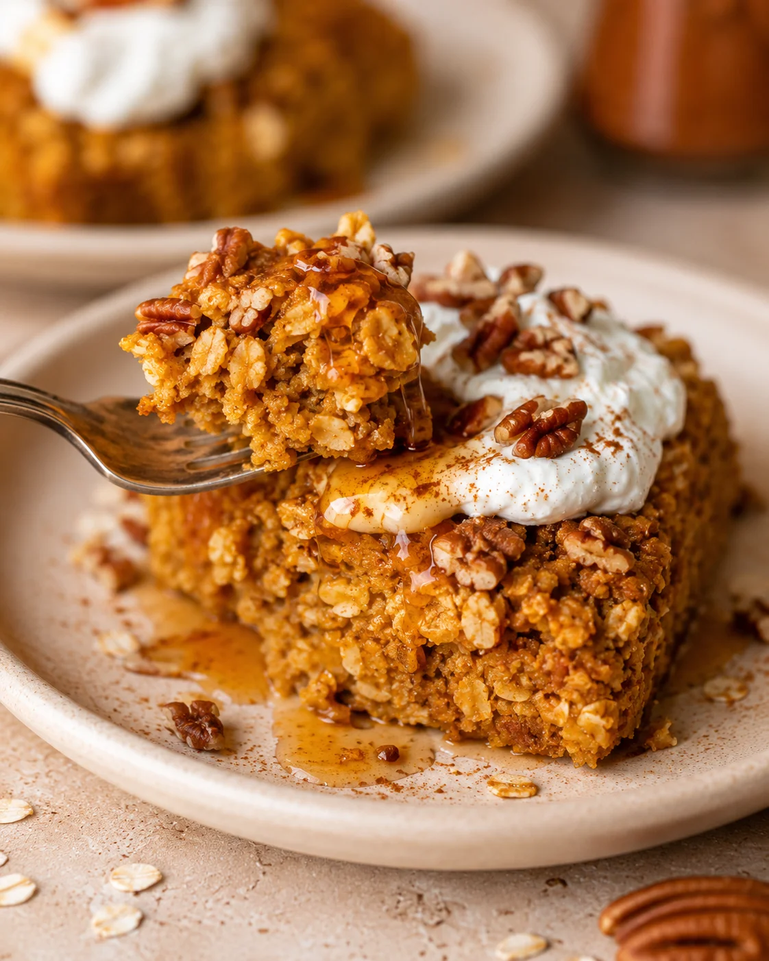 Fork lifting a bite of pumpkin baked oatmeal from a plated square topped with yogurt, pecans, and maple syrup