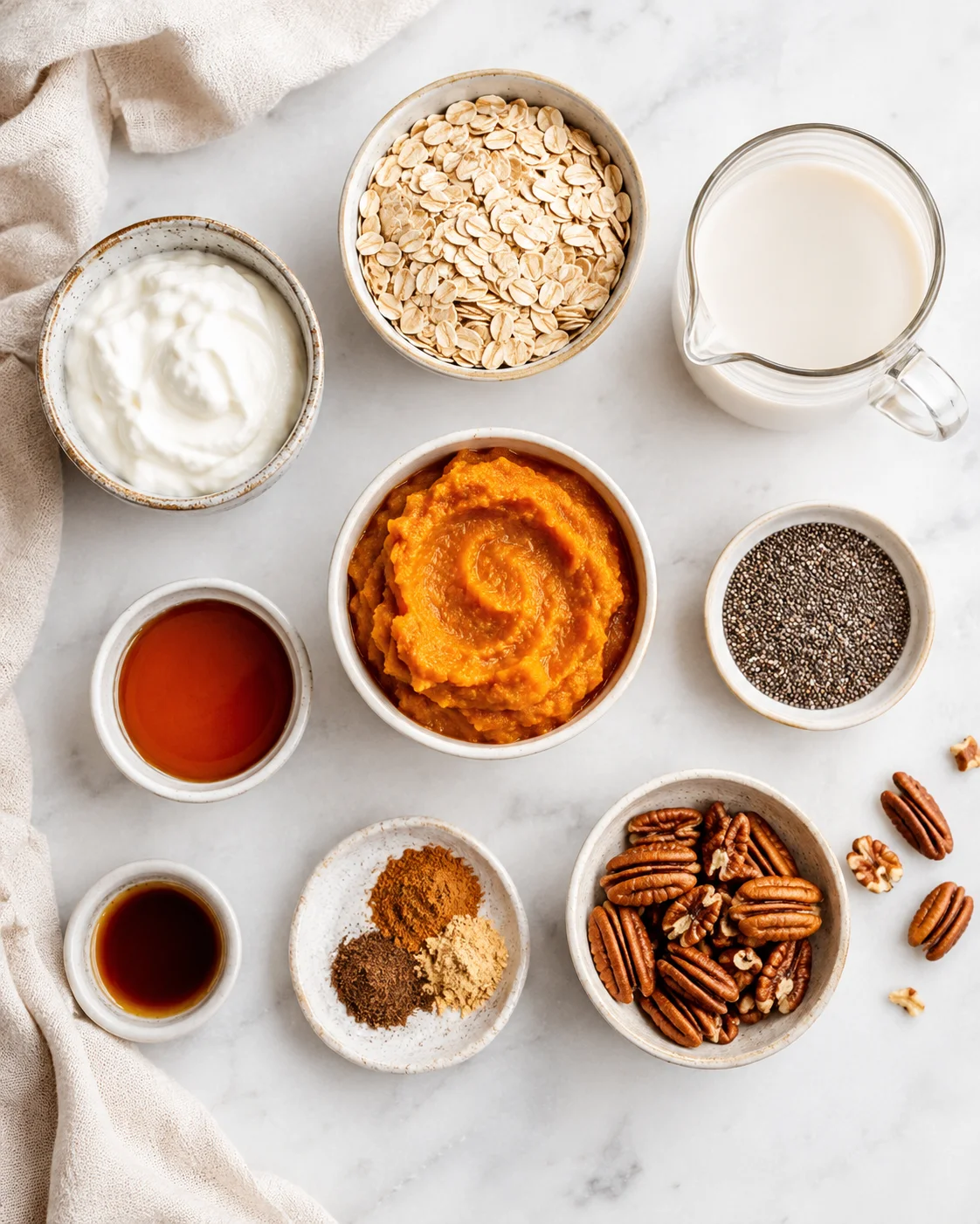 Top-down arrangement of ingredients for pumpkin pie overnight oats including oats, pumpkin puree, yogurt, milk, chia and pecans