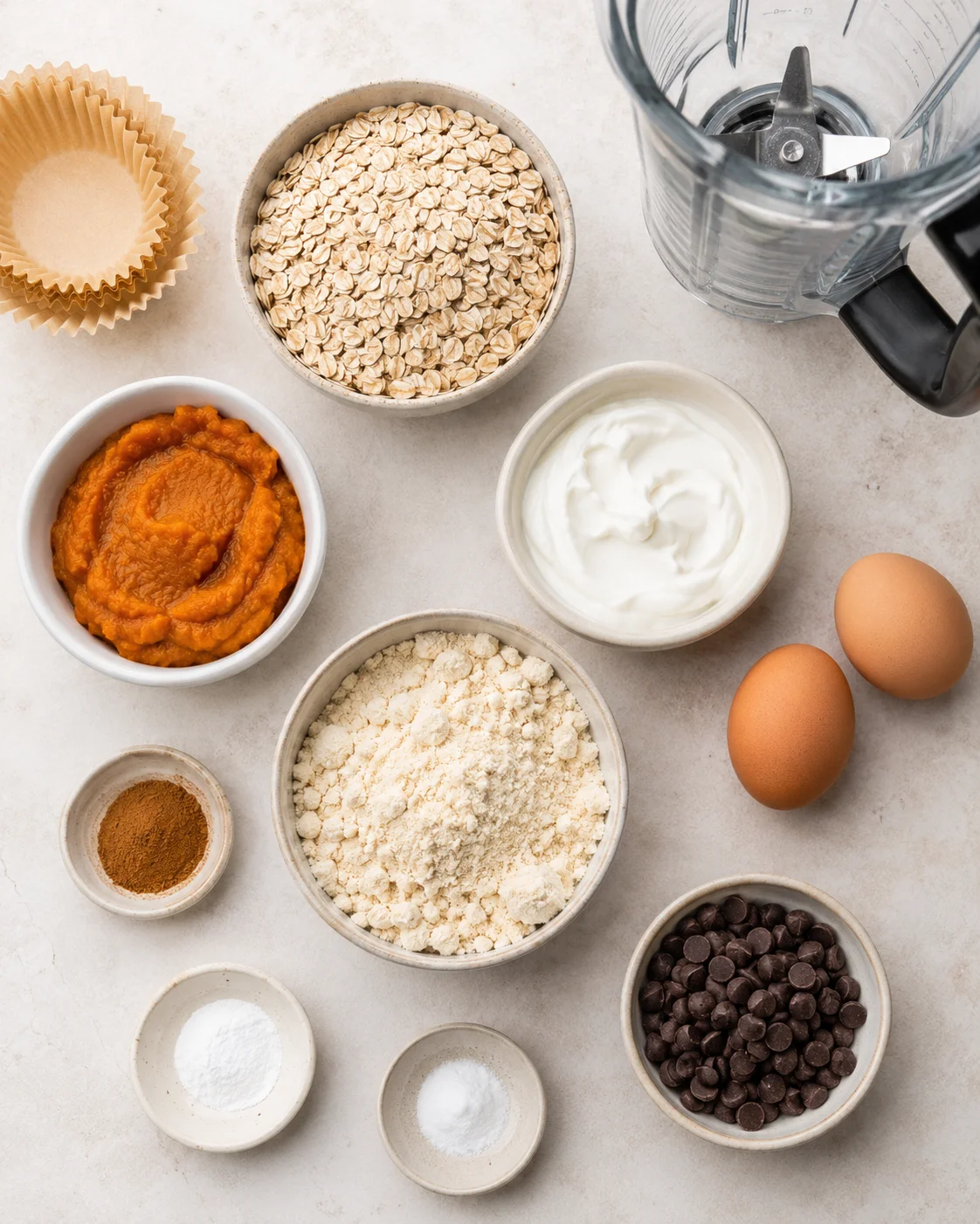 Top-down layout of ingredients for pumpkin chocolate chip protein muffins including oats, pumpkin puree, yogurt, eggs, protein powder and chocolate chips