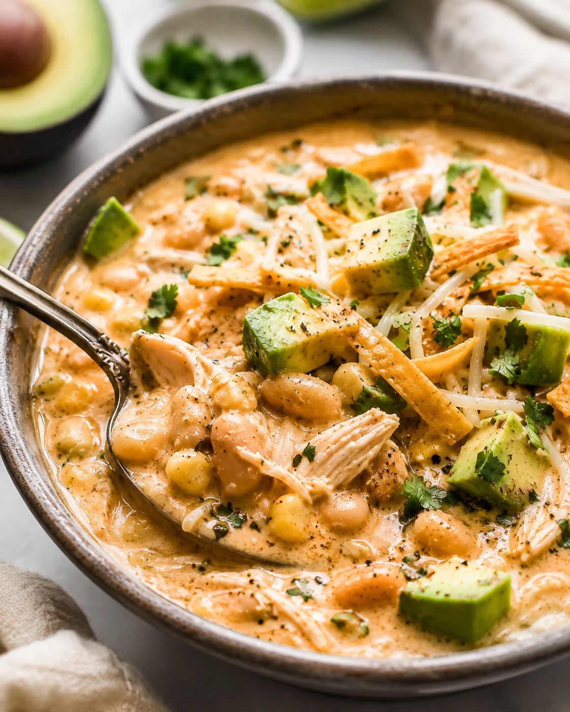 Side view of a bowl of white chicken chili with a spoon and avocado halves in background
